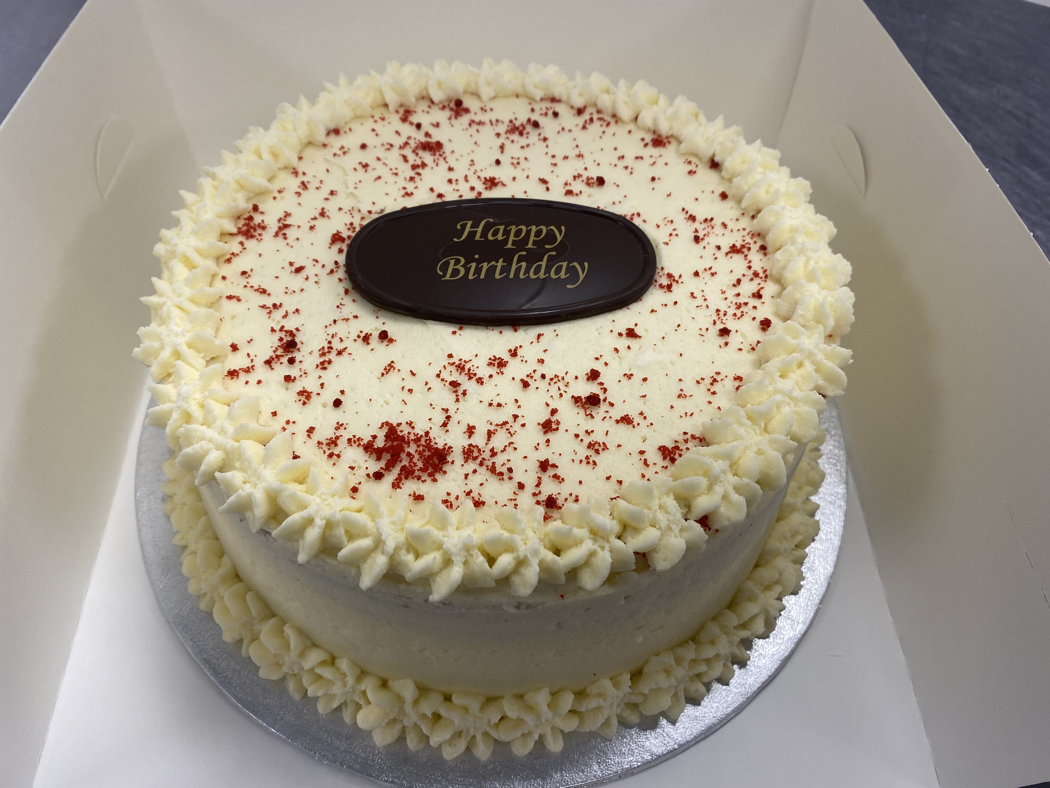 A round red velvet birthday cake topped with red sprinkles and intricate cream cheese frosting along the edges. A chocolate plaque in the center reads 'Happy Birthday.' The cake is placed on a silver board inside a white box, perfect for any special occasion.